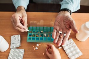 An older adult holding pills in their hand. In the background is a pill organizer on a table